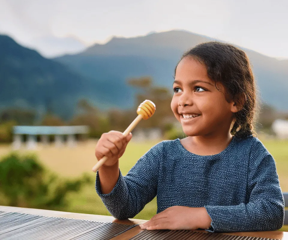 A child at a table in the New Zealand countryside holding a wooden honey dipper